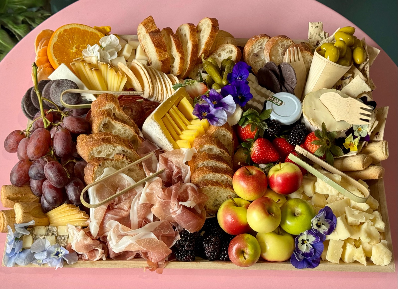 Cheese and charcuterie board with cheeses, prosciutto, baguette, fruit, olives, and edible flowers